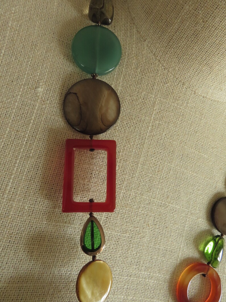 May include: A multi-colored necklace with round, oval, and rectangular beads. The beads are made of various materials, including glass, wood, and metal. The necklace is strung on a brown cord.