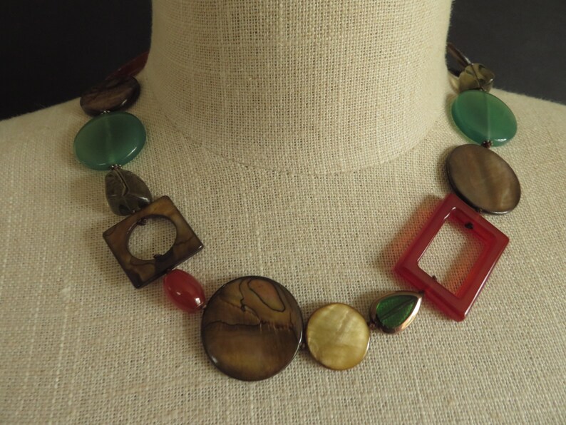 May include: A multi-colored necklace with round and square shaped beads. The beads are made of wood, stone, and glass. The necklace is brown, green, red, and white.