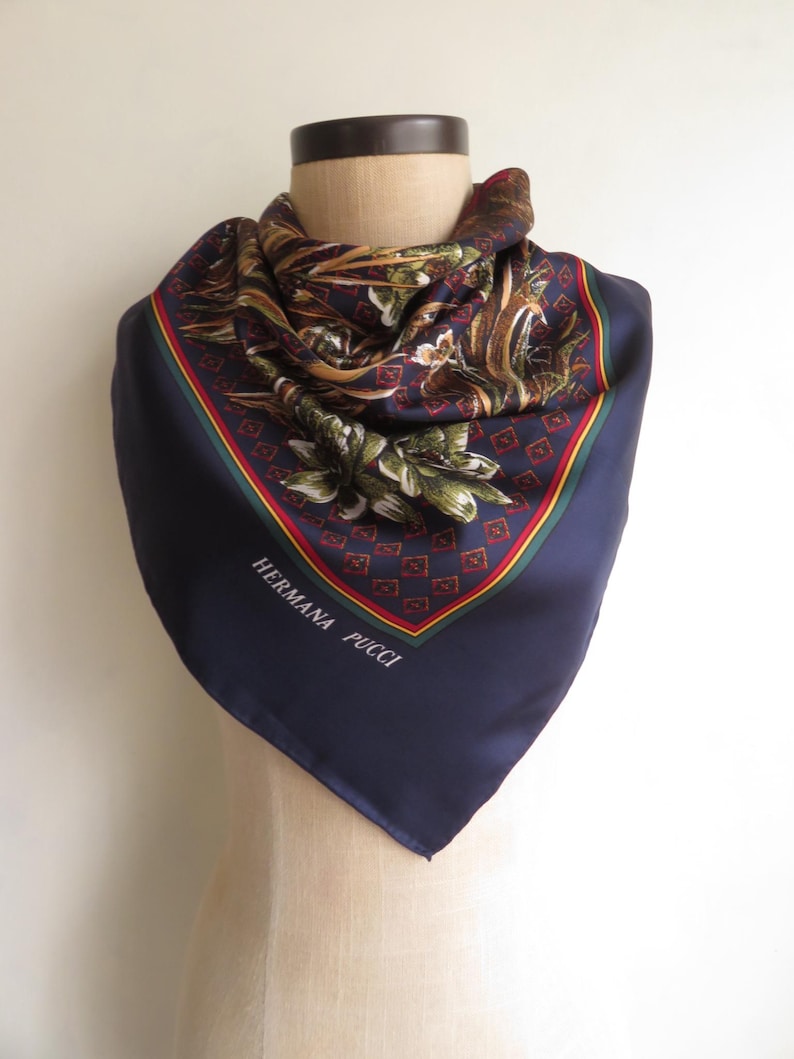 Puede incluir: Un pa&ntilde;uelo de seda azul marino con un estampado floral y geom&eacute;trico. El pa&ntilde;uelo tiene un borde rojo, verde y amarillo y el texto "HERMANA PUCCI" est&aacute; impreso en la esquina. El pa&ntilde;uelo est&aacute; drapeado sobre un maniqu&iacute;.