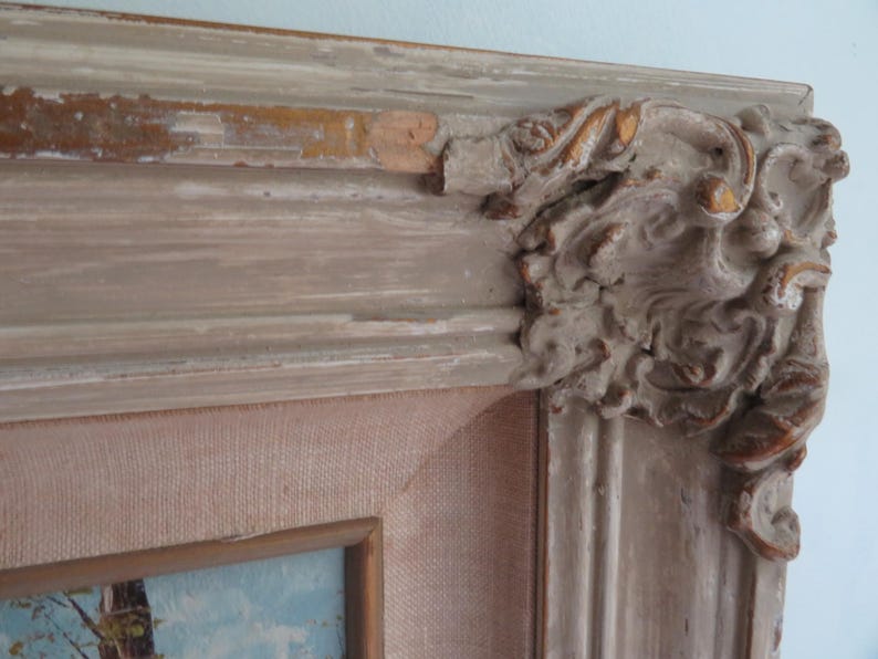 Puede incluir: Primer plano de una esquina de un cuadro enmarcado. El marco es de color blanco roto desgastado con detalles ornamentados y tallados en la esquina. La pintura interior tiene un lienzo rosa claro y un vistazo a un paisaje.