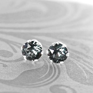 May include: Two gray gemstone stud earrings with a silver setting. The earrings are round with a faceted surface, and are set against a gray background with a subtle pattern. The earrings are a classic jewelry item.