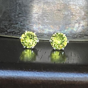 May include: A pair of round, light green gemstone stud earrings. The earrings are set in a silver metal. The gemstones have a faceted cut, and the earrings are displayed on a dark wooden surface.