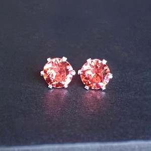 Peut inclure: Deux boucles d'oreilles à clous avec des pierres précieuses rondes de couleur pêche serties dans du métal argenté. Les boucles d'oreilles sont présentées sur une surface sombre et texturée, contrastant avec la couleur et l'éclat des pierres.