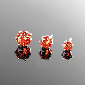 Op de afbeelding: Drie sieraden met oranjerode edelstenen. De edelstenen zijn gezet in een zilverkleurig metaal, met een reflecterend oppervlak eronder. De maten van de edelstenen variëren, met de grootste links en de kleinste rechts.
