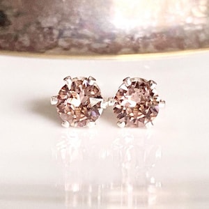 May include: A pair of stud earrings with light brown gemstones set in silver. The earrings are round and faceted, reflecting light. The background is a blurred, metallic surface and a white surface.