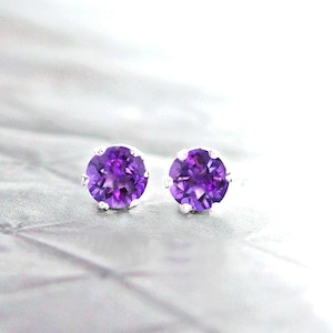 May include: A pair of stud earrings featuring round, vibrant purple gemstones set in silver. The gemstones are a deep violet color, and the earrings are displayed on a light, textured surface.