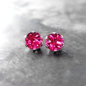 May include: A pair of stud earrings featuring vibrant, hot pink gemstones set in a silver metal. The round-cut stones are the focal point, catching the light and creating a sparkling effect. The earrings are displayed on a textured, gray surface.
