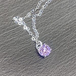 May include: A silver necklace with a round, light purple gemstone pendant. The pendant is set in a silver prong setting and hangs from a delicate silver chain. The necklace is displayed on a textured gray surface.