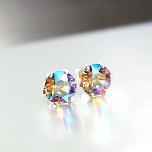 May include: A pair of iridescent stud earrings. Each earring features a round, multi-colored gemstone set in a silver-toned, pronged setting. The gemstones display hues of blue, gold, and purple, reflecting light. The earrings are displayed on a reflective, silver surface.