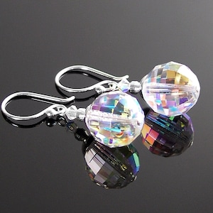 May include: A pair of iridescent crystal earrings with faceted, spherical beads. The earrings have silver-colored hooks and small accent beads. The crystals reflect a rainbow of colors, creating a sparkling effect. The earrings are displayed on a reflective black surface.