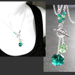 Puede incluir: Un collar de plata con cierre de palanca y dos colgantes de cristal verde. Los colgantes tienen forma de tr&eacute;bol de cuatro hojas. El collar est&aacute; en un maniqu&iacute; blanco que lleva una camiseta sin mangas negra.