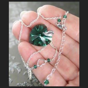May include: A silver chain necklace with a green crystal heart pendant and a small silver leaf charm. The necklace has small green crystal accents.