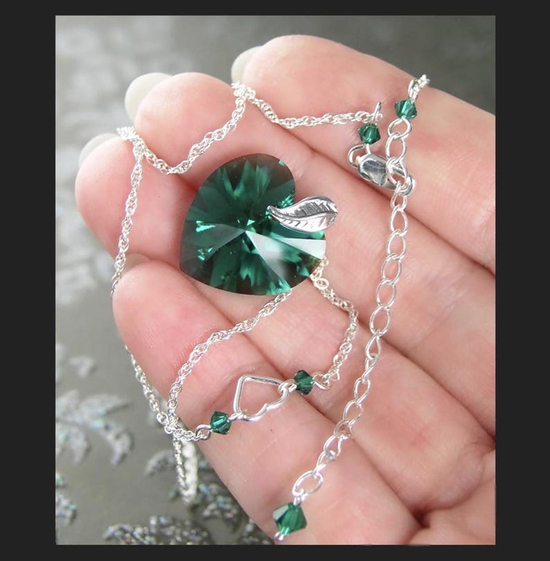 May include: A silver chain necklace with a green crystal heart pendant and a small silver leaf charm. The necklace has small green crystal accents.