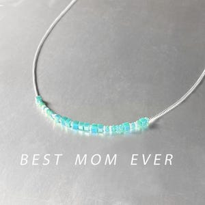 Op de afbeelding: Een zilveren ketting met een delicate rij lichtblauwe en witte kralen. De ketting heeft de tekst "BEST MOM EVER" eronder gedrukt.