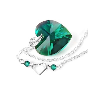 May include: A silver necklace with a large emerald green heart-shaped crystal pendant. The pendant has a leaf design on the bail. The necklace also has a smaller heart charm and emerald green beads.