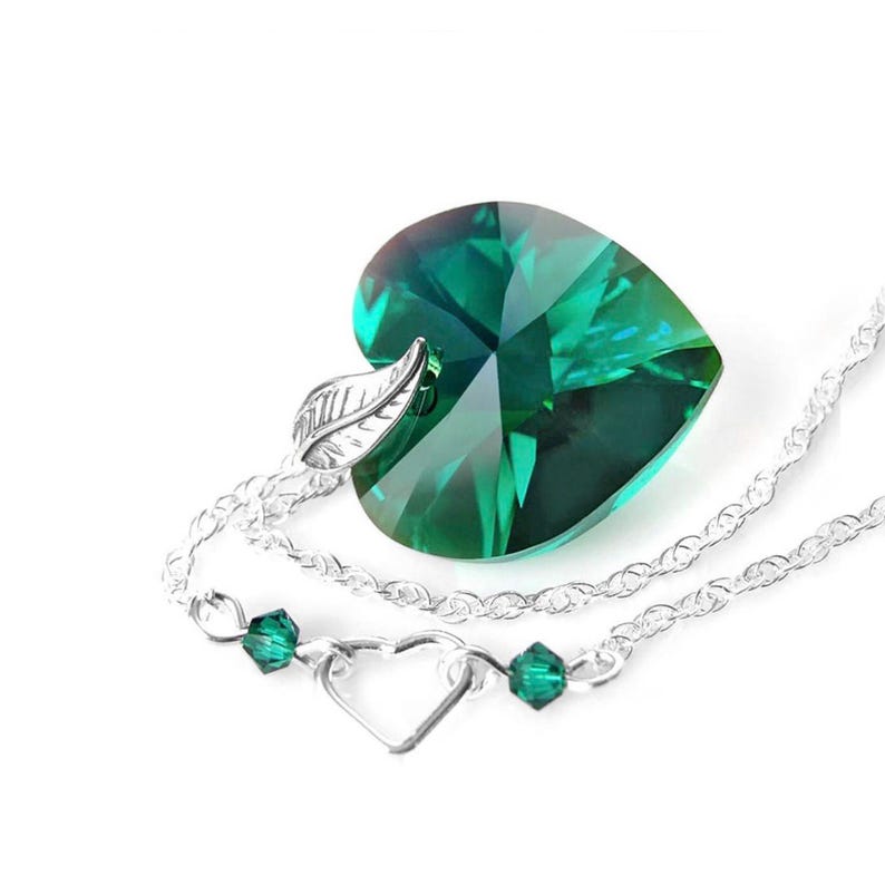 May include: A silver necklace with a large emerald green heart-shaped crystal pendant. The pendant has a leaf design on the bail. The necklace also has a smaller heart charm and emerald green beads.