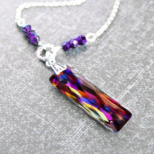 May include: A silver chain necklace with a purple crystal pendant. The pendant is a long, rectangular prism with a faceted, iridescent finish that shows shades of red, orange, yellow, blue, and purple.