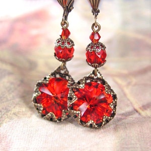 May include: A pair of ornate, vintage-style earrings. Each earring features a large, faceted red crystal pendant with a decorative bronze-colored metal frame. Smaller red crystals and bronze accents complete the design, creating a striking, elegant look.