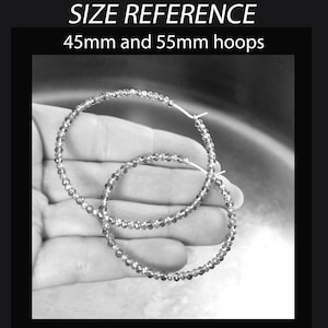May include: Two silver hoop earrings, one 45mm and one 55mm in diameter, are shown in a hand. The earrings are made of small, round beads.