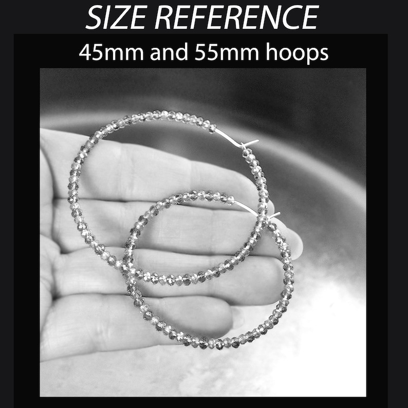 May include: Two silver hoop earrings, one 45mm and one 55mm in diameter, are shown in a hand. The earrings are made of small, round beads.