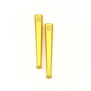May include: Two gold-colored, cone-shaped objects with a sunburst design etched near the top. The objects have a rounded base and are set against a white background. The objects appear to be decorative or functional.