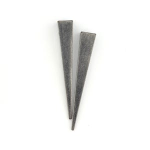 May include: A pair of silver-coloured, triangular earrings with a matte finish. The earrings are long and slender, tapering to a point. They are set against a white background.