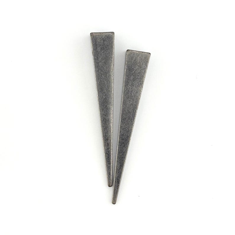 May include: A pair of silver-coloured, triangular earrings with a matte finish. The earrings are long and slender, tapering to a point. They are set against a white background.
