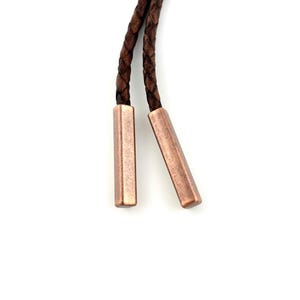 May include: Close-up of a bolo tie featuring a brown braided leather cord and two copper-colored rectangular metal tips. The tips are angled slightly away from each other. The background is white.