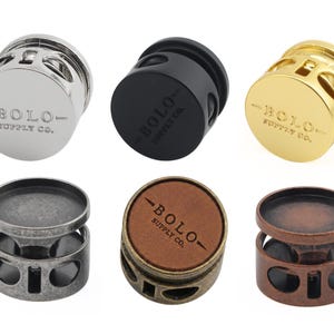 May include: Six cylindrical bolo tie slides featuring cut-out side details. The top row includes polished silver, matte black, and gold finishes. The bottom row features antique silver, a brown leather-topped bronze, and copper, all stamped with BOLO SUPPLY CO.