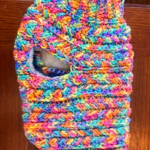 May include: A vibrant, hand-knitted balaclava in a rainbow of colors, including blue, orange, yellow, and purple. The balaclava features a face opening and a soft, textured appearance. This cozy headwear is perfect for cold weather.