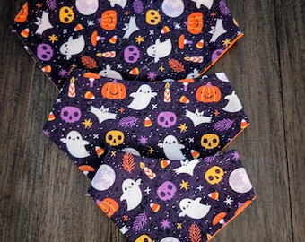 Puede incluir: Tres baberos con temática de Halloween con un fondo morado oscuro y un borde naranja. Los baberos presentan un patrón repetido de fantasmas blancos, calabazas naranjas, calaveras moradas y caramelos de maíz. Los baberos están hechos de tela suave.