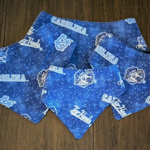May include: Blue dog bandanas with a repeating pattern of the word "Carolina", the Tar Heels logo, and the UNC logo. The bandanas are in various sizes and are made of a blue fabric.