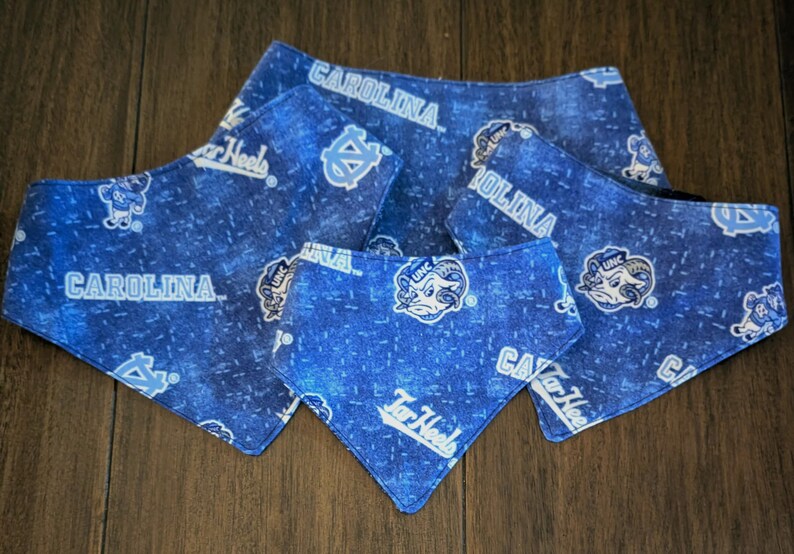 May include: Blue dog bandanas with a repeating pattern of the word "Carolina", the Tar Heels logo, and the UNC logo. The bandanas are in various sizes and are made of a blue fabric.
