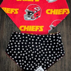 May include: Two dog bandanas. The top bandana is red with yellow text that says "CHIEFS" and a helmet graphic. The bottom bandana is black with white paw prints. A tag reads "Handmade by Wigglebutts NC".