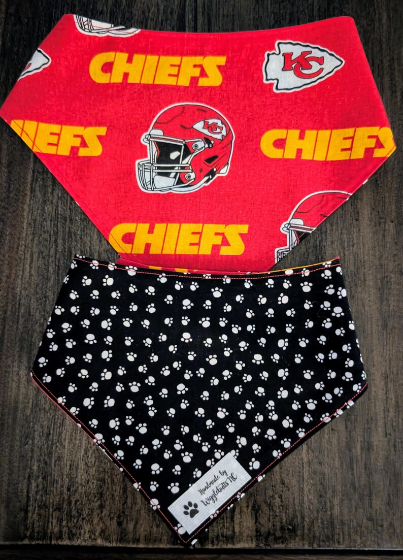 May include: Two dog bandanas. The top bandana is red with yellow text that says "CHIEFS" and a helmet graphic. The bottom bandana is black with white paw prints. A tag reads "Handmade by Wigglebutts NC".