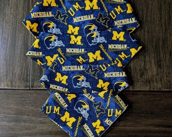 May include: A stack of four blue and yellow patterned bandanas. The bandanas feature the text "MICHIGAN" and "UM" along with a graphic of a football helmet. The bandanas are made of fabric.