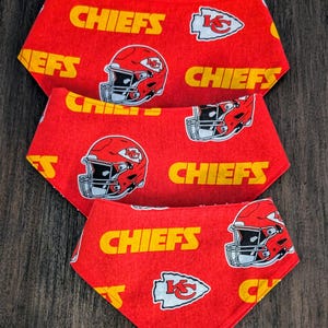 May include: Three red pet bandanas with a repeating pattern of yellow "CHIEFS" text, football helmets, and team logos. The bandanas are arranged in a layered, triangular shape.