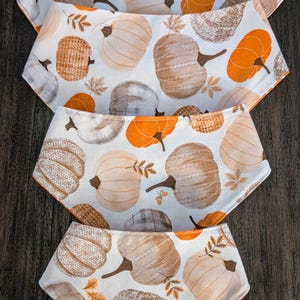 Puede incluir: Cuatro baberos para bebé con un estampado de calabazas naranjas, marrones y grises sobre fondo blanco. Los baberos tienen un ribete naranja y están apilados verticalmente. Hechos de tela.