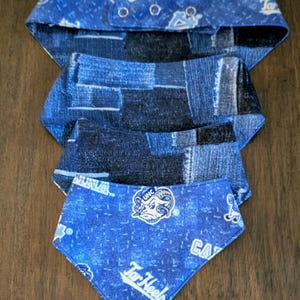 May include: Four blue dog bandanas. The top bandana has a denim pattern with snap closures. The second and third bandanas have a denim patchwork pattern. The bottom bandana has a blue background with the text "Tar Heels" and a ram logo.