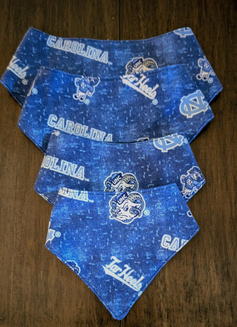 May include: Four blue dog bandanas with a repeating pattern of the words "Carolina", "Tar Heels", and the UNC logo. The bandanas are in various sizes and are made of a soft fabric.
