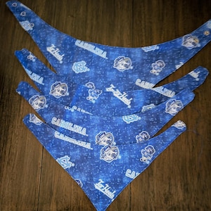 May include: Four blue dog bandanas with a repeating pattern of the words "CAROLINA" and "Tar Heels", along with the university's logo. The bandanas are made of fabric and have a snap closure.