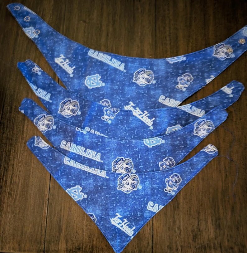 May include: Four blue dog bandanas with a repeating pattern of the words "CAROLINA" and "Tar Heels", along with the university's logo. The bandanas are made of fabric and have a snap closure.