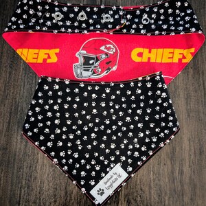 May include: Dog bandana featuring a red and black design. The top section has a black background with white paw prints and a red band with a football helmet graphic and the word "CHIEFS". The bottom section is black with white paw prints.