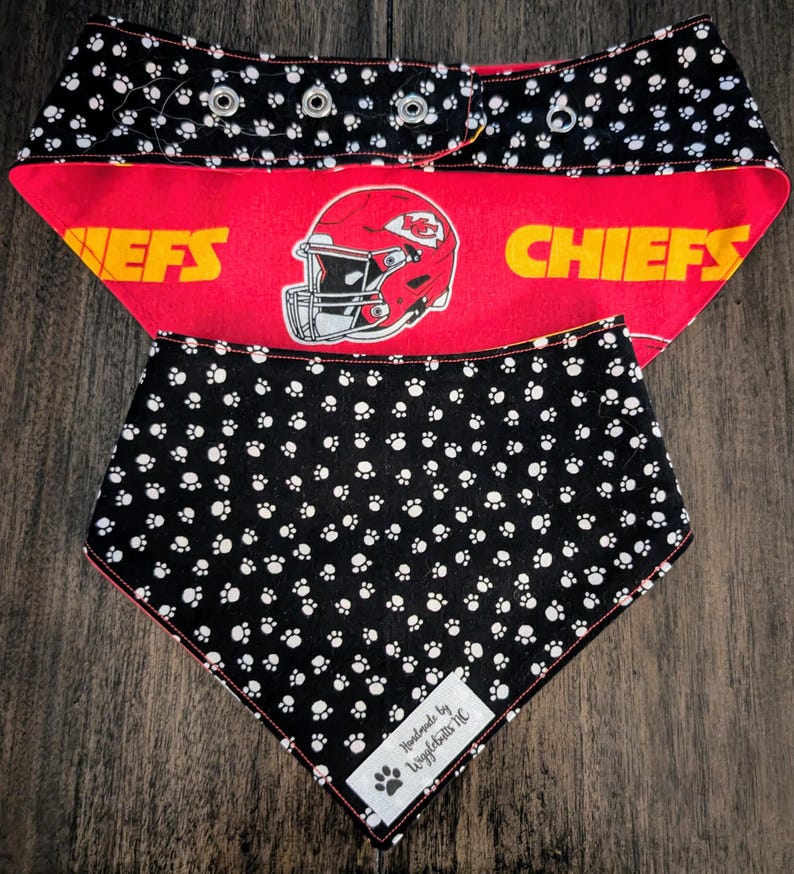 May include: Dog bandana featuring a red and black design. The top section has a black background with white paw prints and a red band with a football helmet graphic and the word "CHIEFS". The bottom section is black with white paw prints.