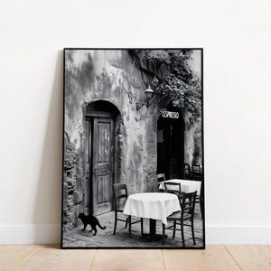 Op de afbeelding: Zwart-witfoto van een café in Italië. Het café heeft een stenen muur met een houten deur en een bord met de tekst "Espresso". Er staan twee tafels met witte tafelkleden en stoelen buiten het café. Een zwarte kat loopt voor het café.