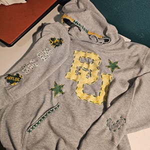 Peut inclure: Sweat-shirt à capuche gris avec un grand "BU" brodé en jaune sur le devant. Le sweat-shirt a des étoiles vertes et jaunes et d'autres détails brodés sur les manches et les poches.