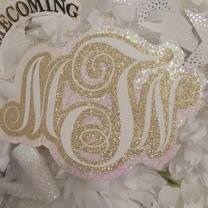 May include: A decorative monogram with the letters "MN" in gold glitter, set against a white and pink background. The word "HOMECOMING" is visible in gold lettering above the monogram. The background is white.