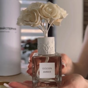 May include: A square glass fragrance diffuser bottle filled with amber-colored liquid. A white label on the front reads GOLDEN AMBER. The bottle features a white cylindrical cap with embossed botanical details and a bouquet of white flower-shaped reeds.
