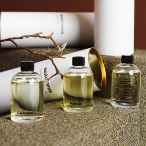 Pode incluir: Três frascos de vidro transparente com tampas pretas contendo líquido claro, rotulados como Caramel, Mango e Wine. Cada frasco é acompanhado por um tubo cilíndrico de papelão branco com o texto MAЙCTEPНЯ impresso em preto.