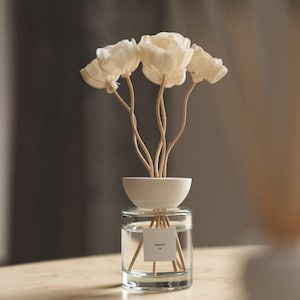 Pode incluir: Um difusor de varetas em vidro transparente com um topo de cerâmica branca em forma de taça e quatro flores de madeira na cor creme. Um pequeno rótulo quadrado branco com texto preto está centralizado no recipiente de vidro.